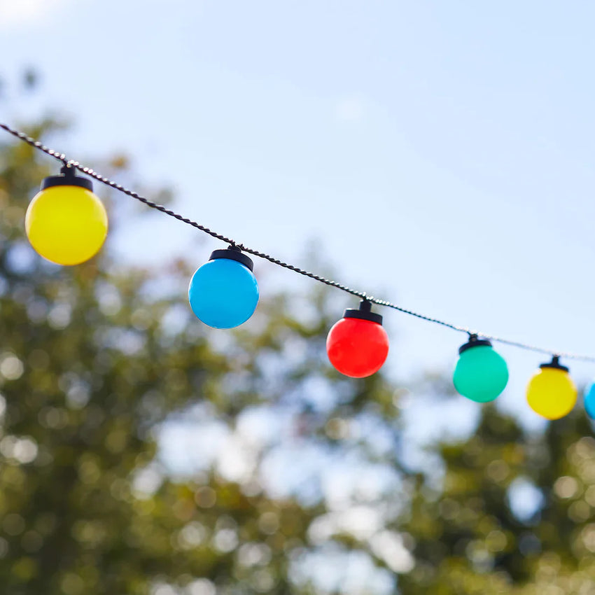 Bunte Outdoor-Lichterkette mit gelben, blauen, roten und grünen Kugelleuchten vor unscharfem Gartenhintergrund