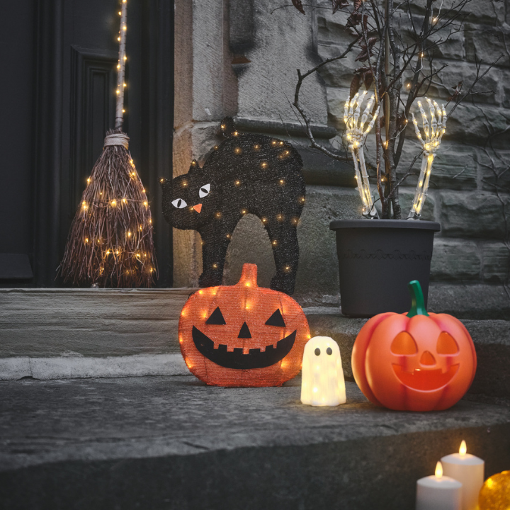 Halloween-Deko mit leuchtenden Kürbissen, Geist, Katze, Hexenbesen und Kerzen vor einer Haustür.