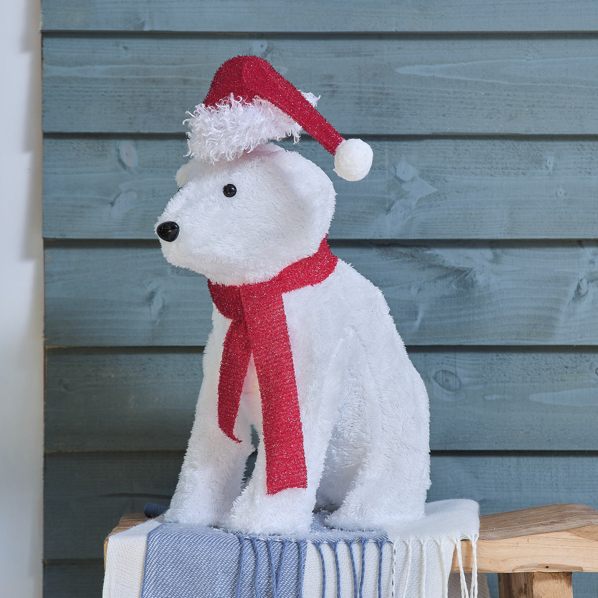 Süße weiße Plüsch-Tierfigur mit roter gestrickter Weihnachtsmütze und rotem Schal-Detail, positioniert auf blauem Holzuntergrund – Premium-Plüsch-Dekofigur für detailreiche Weihnachtsdisplays und gehobene festliche Raumdekorationen mit charaktervollem Design.